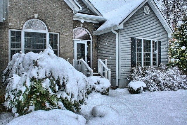 A brick-and-vinyl-siding home with bushes in front, all covered in several inches of snow.