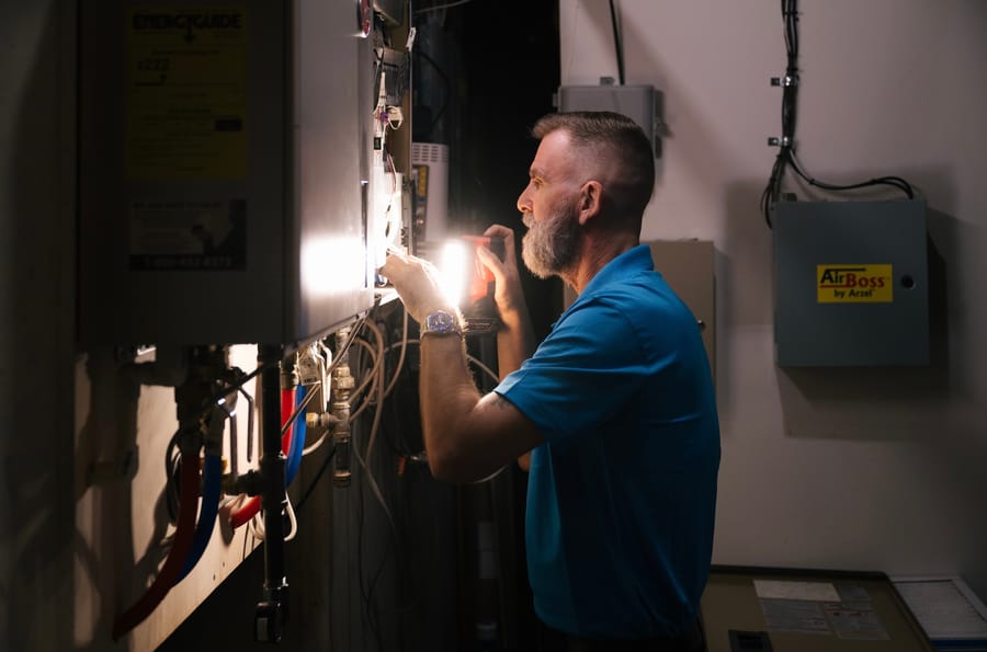 An HVAC technician in a blue shirt inspects an electrical panel with a flashlight, surrounded by cables and HVAC equipment in a dimly lit space.