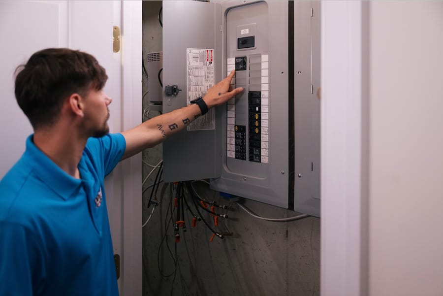 A technician in a blue shirt adjusts a residential electrical panel, showcasing maintenance work in a utility space.
