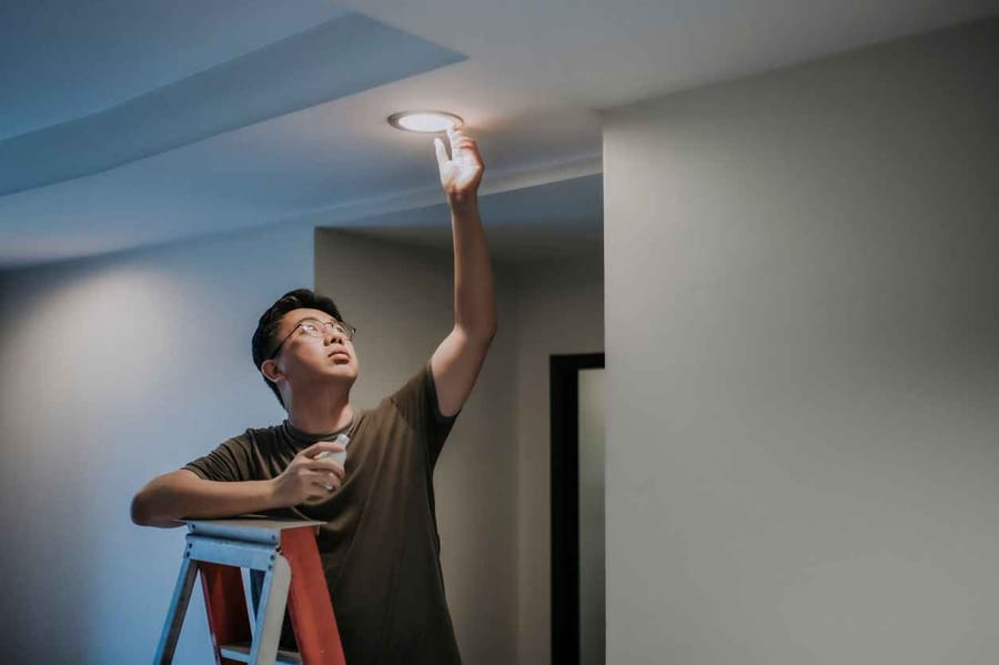 An adult male standing on a ladder in his living room changing a light bulb